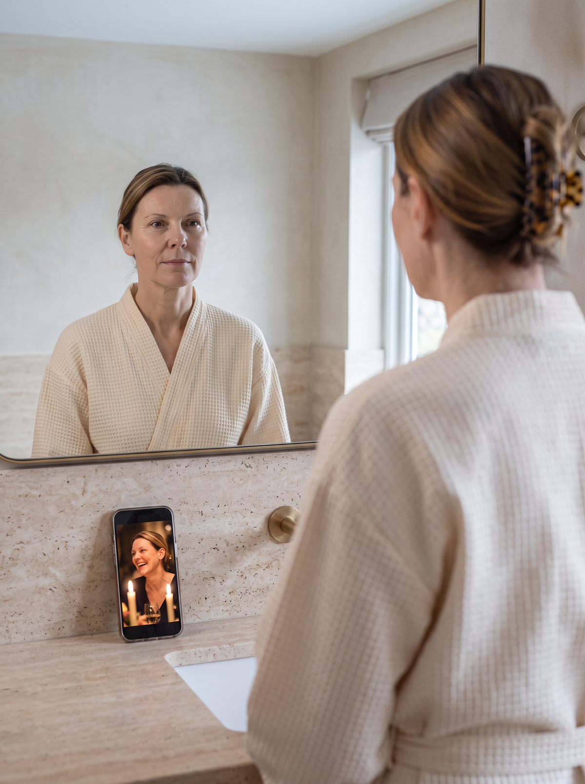 Le matin, une femme se regarde de face dans le miroir ; son téléphone posé sur le lavabo affiche une photo d’elle prise lors d’un dîner